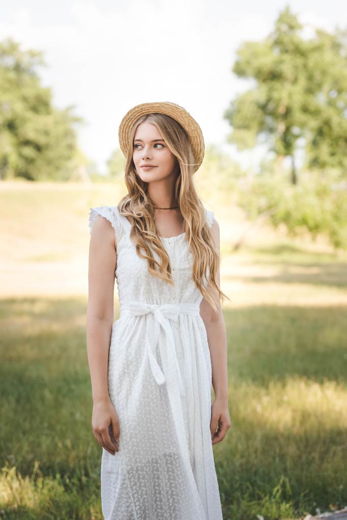 mooi meisje met stro hoed en witte jurk staande op weide en op zoek weg - Foto, afbeelding