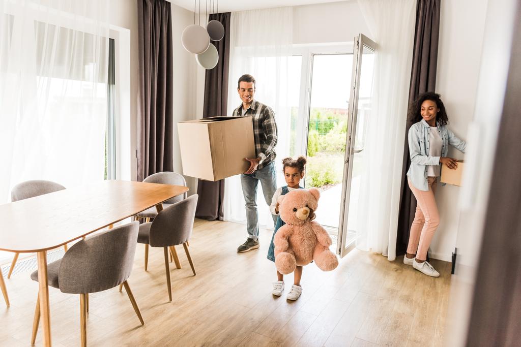 täyspitkä näkymä afrikkalainen amerikkalainen perhe kävelee taloon, kun vanhemmat tilalla laatikot ja lapsi tilalla nallekarhu
  - Valokuva, kuva