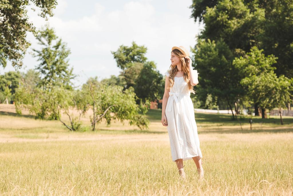 schönes Mädchen in weißem Kleid und Strohhut steht auf der Wiese und schaut weg - Foto, Bild