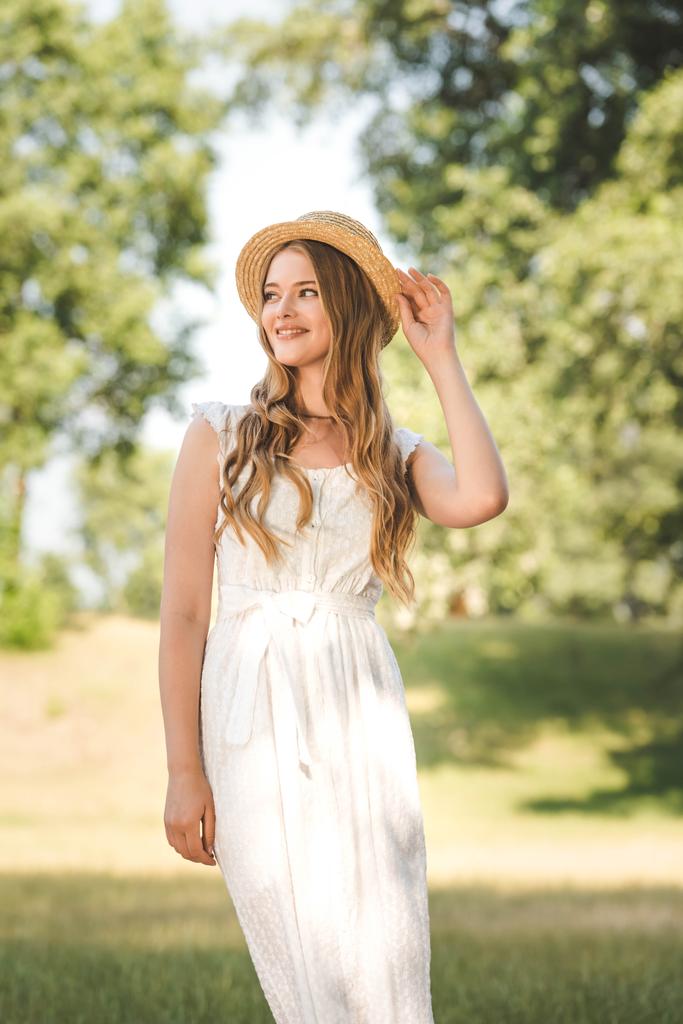 piękna dziewczyna w białej sukni dotykając słomy kapelusz stojąc na łące i patrząc od - Zdjęcie, obraz
