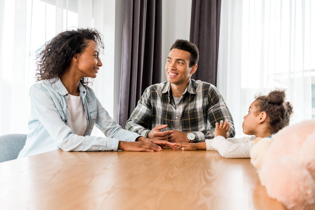 afrikanisch-amerikanische Familie sitzt vor Tisch und lächelt, während sich die Eltern anschauen - Foto, Bild