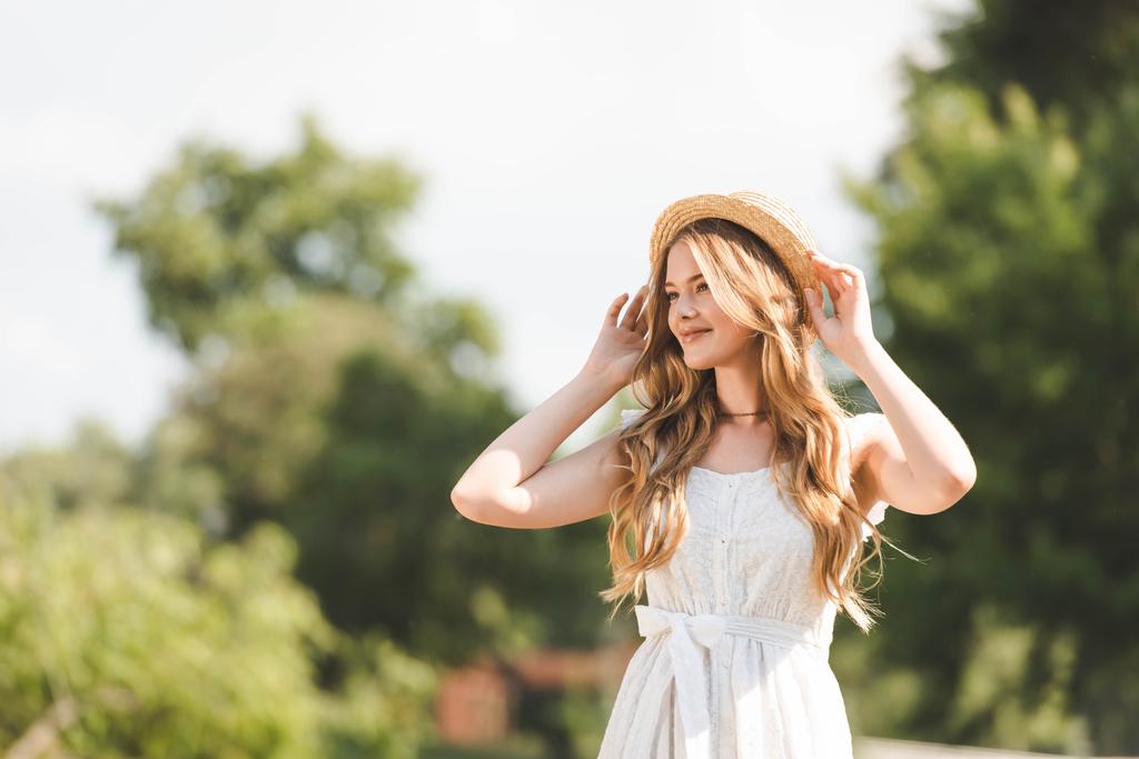 menina bonita em vestido branco tocando chapéu de palha e sorrindo enquanto estava em pé no prado e olhando para longe
 - Foto, Imagem