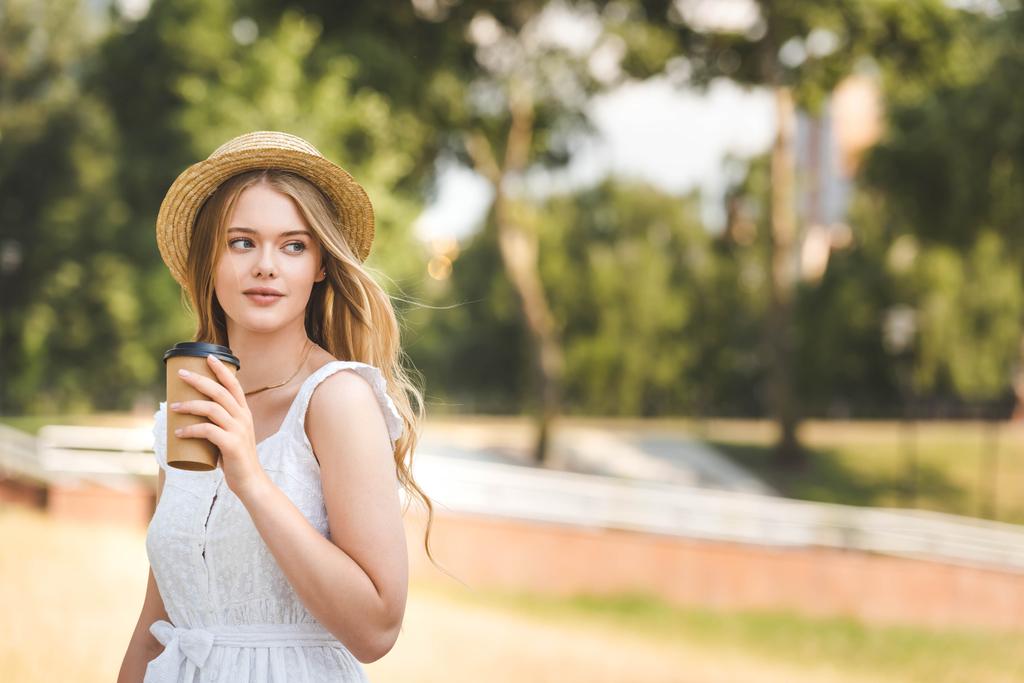 schönes Mädchen in weißem Kleid und Strohhut hält Kaffeetasse aus Papier und schaut weg - Foto, Bild