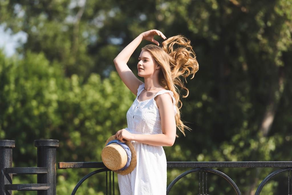 piękna dziewczyna w białej sukni trzymając słomiany kapelusz podczas dotykania włosów i patrząc z dala - Zdjęcie, obraz