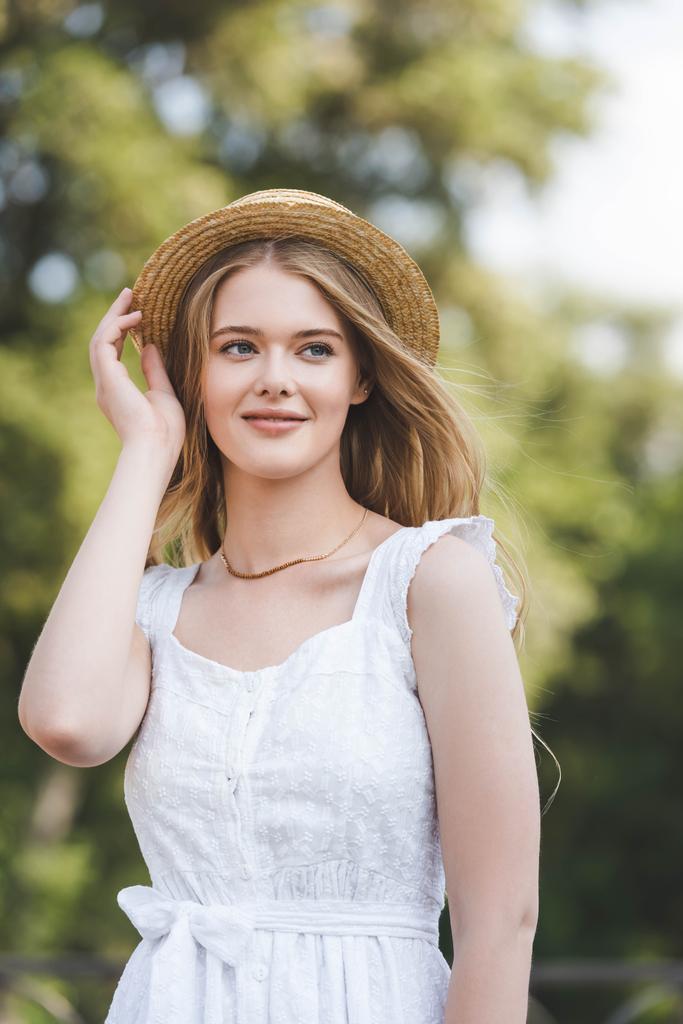 vista frontal de la hermosa joven en vestido blanco tocando sombrero de paja mientras sonríe y mira hacia otro lado
 - Foto, Imagen