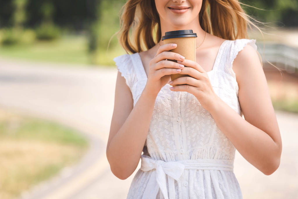 обрезанный вид на молодую девушку в белом платье и соломенной шляпе улыбаясь и держа бумажный кофе чашку
  - Фото, изображение