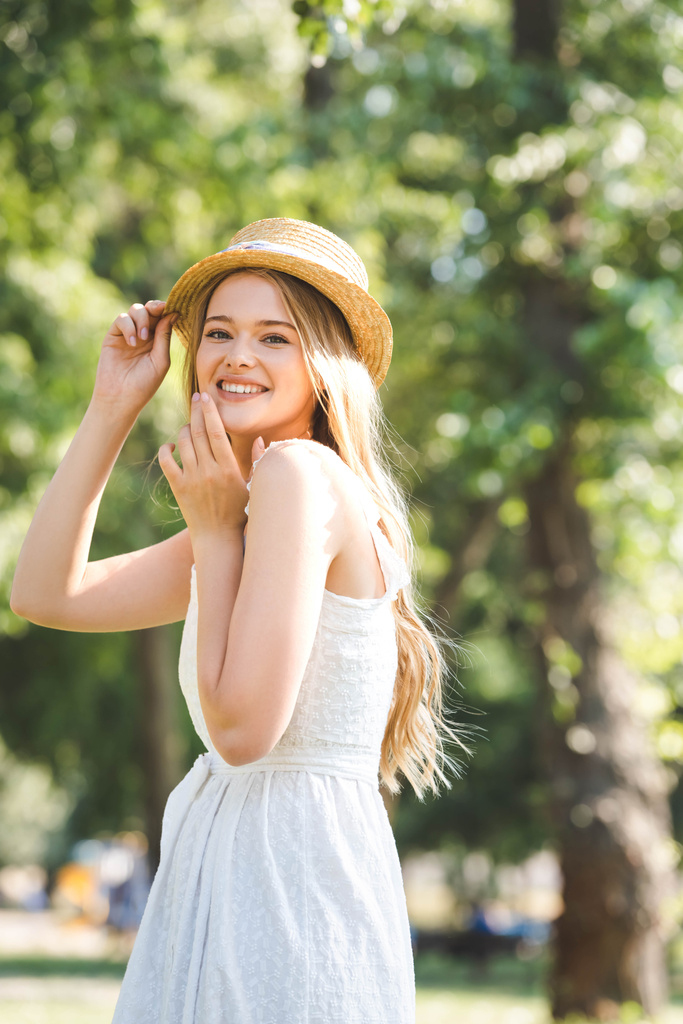 白いドレスと麦わら帽子の美しい若い女性の選択的な焦点は、笑顔とカメラを見て - 写真・画像