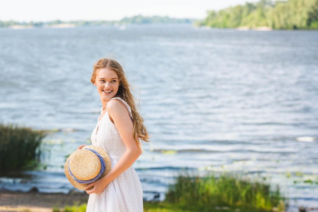 giovane ragazza in piedi sulla riva del fiume, mentre tenendo cappello di paglia e guardando altrove
 - Foto, immagini