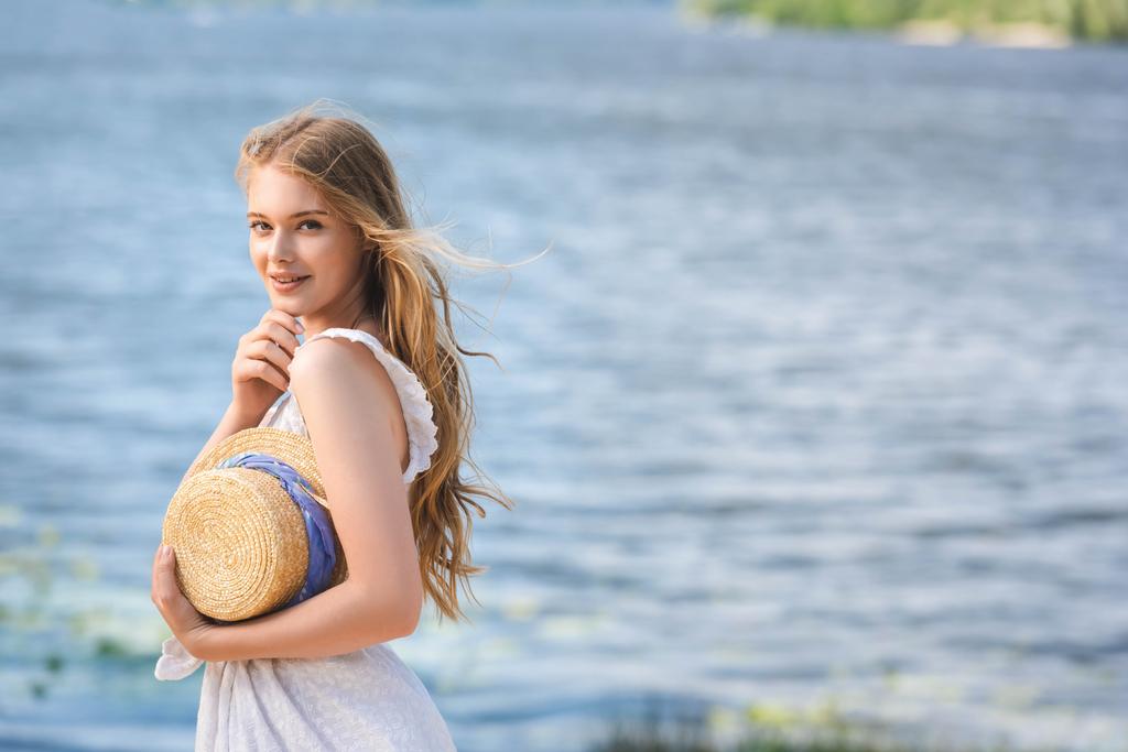 beautiful young girl standing on shore of river while holding straw hat and looking at camera - Photo, Image