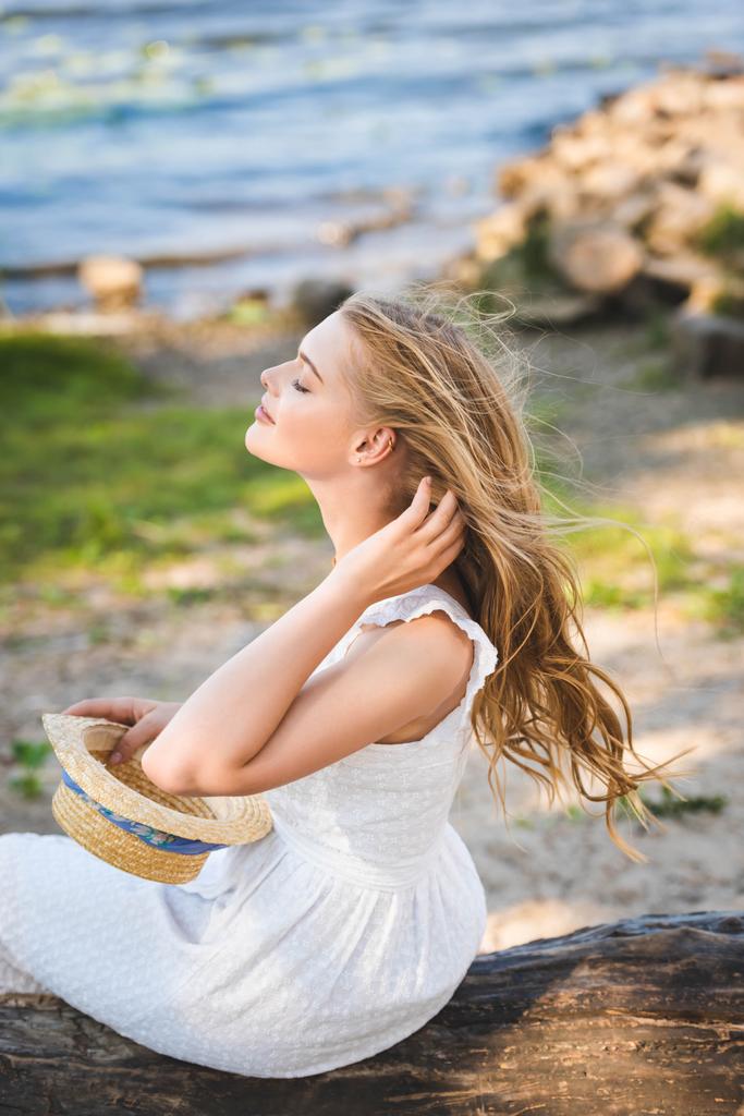 όμορφο κορίτσι σε λευκό φόρεμα κρατώντας ψάθινο καπέλο ενώ καθιστός ξύλινος κορμός με κλειστά μάτια - Φωτογραφία, εικόνα