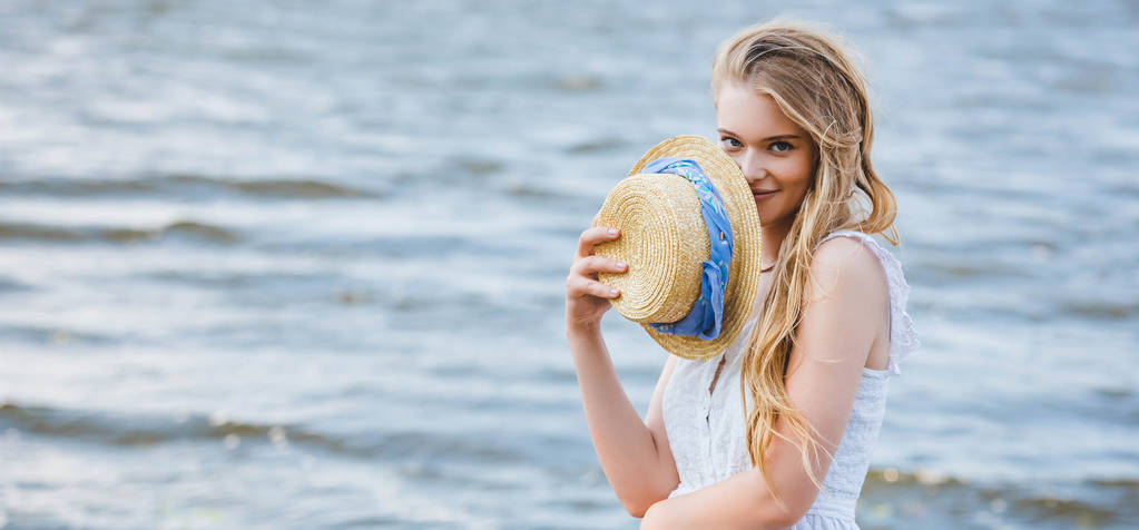 πανοραμικό σφηνάκι όμορφο κορίτσι κρατώντας ψάθινο καπέλο κοντά στο πρόσωπο, χαμογελώντας και κοιτάζοντας την κάμερα - Φωτογραφία, εικόνα