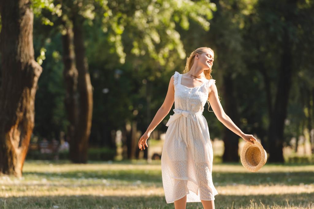 mooi meisje in witte jurk houden stro hoed terwijl glimlachend en staan op weide met gesloten ogen - Foto, afbeelding