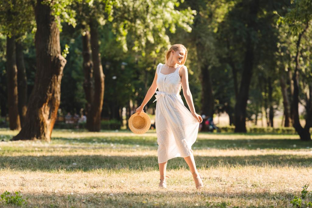 full length view of beautiful girl in white dress holding straw hat while smiling and standing on meadow with closed eyes - Photo, Image