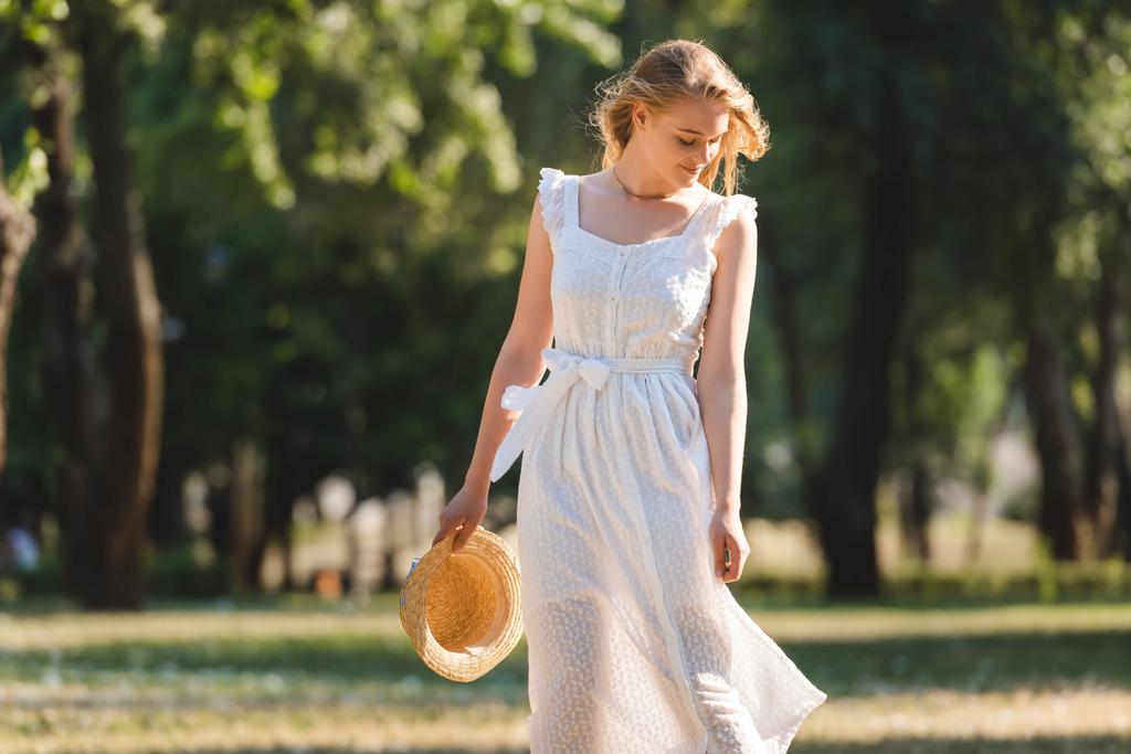 menina bonita em vestido branco segurando chapéu de palha enquanto sorrindo e de pé no prado e olhando para baixo
 - Foto, Imagem