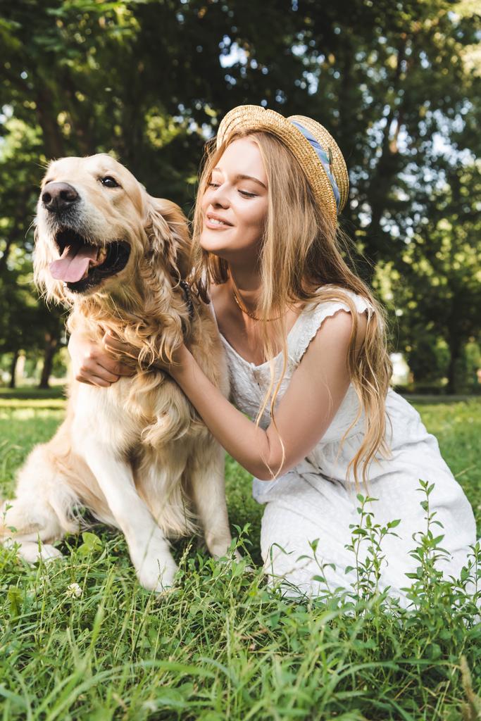 beautiful girl in white dress and straw hat petting golden retriever while smiling, sitting on meadow and looking at dog - Photo, Image