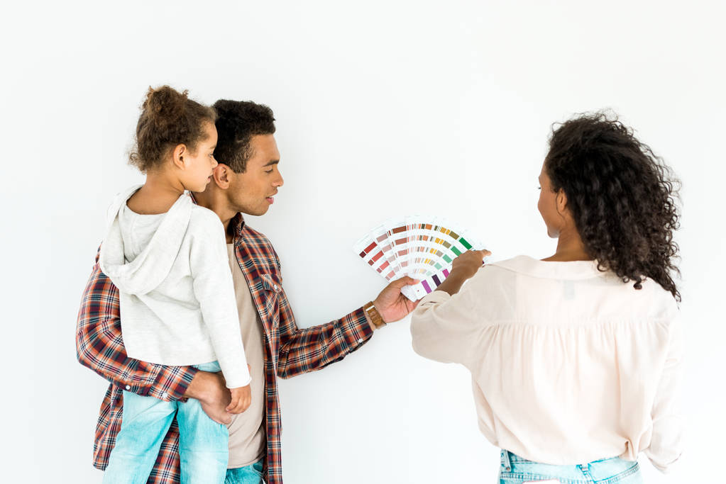 African American ojciec i matka próbuje wybrać kolor ich ściany z kolorowej palety, podczas gdy człowiek trzyma dziecko na białym tle - Zdjęcie, obraz