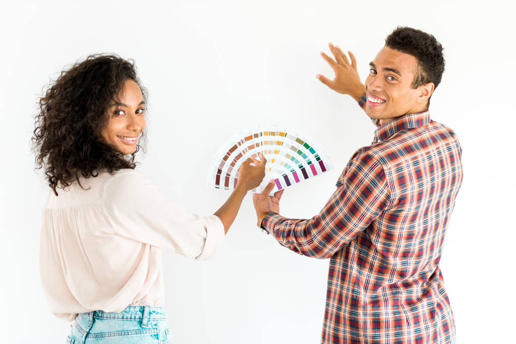 Αφρικανός Αμερικανός άνδρας και γυναίκα προσπαθεί να επιλέξει το χρώμα για τον τοίχο τους από πολύχρωμη παλέτα και κοιτάζοντας την κάμερα απομονώνεται σε λευκό - Φωτογραφία, εικόνα