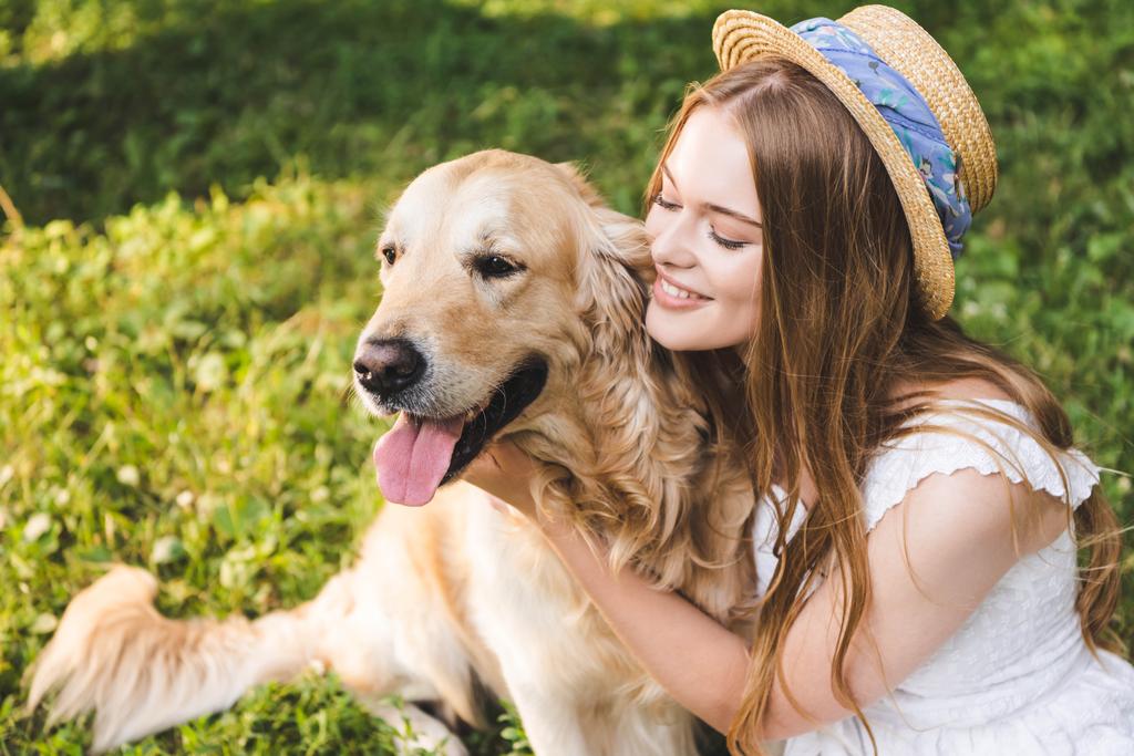 mooi meisje in witte jurk en stro hoed knuffelen Golden Retriever terwijl zittend op weide en kijken naar hond - Foto, afbeelding
