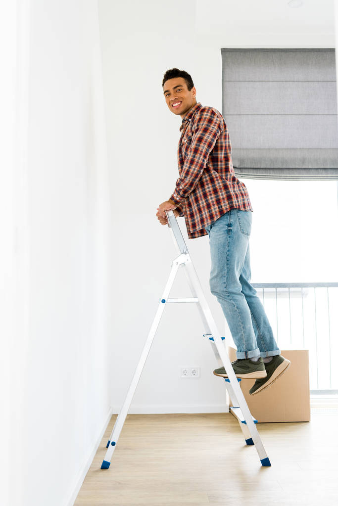 zobrazení plné délky afrického amerického muže stojícího na žebříku a pohledu na kameru  - Fotografie, Obrázek