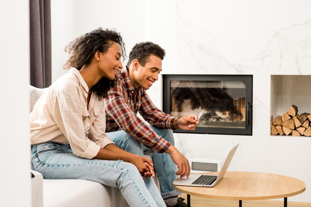 vista laterale della coppia seduta sul divano e guardando il computer portatile mentre l'uomo in possesso di carta di credito
  - Foto, immagini