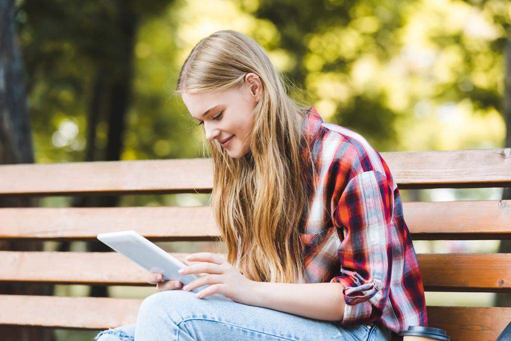 parkta ahşap bankta oturan ve dijital tablet kullanarak rahat giysiler içinde güzel kız seçici odak - Fotoğraf, Görsel