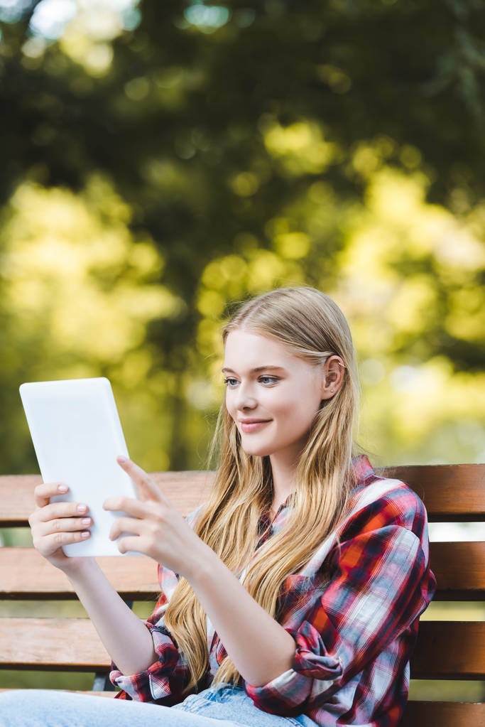 parkta ahşap bankta oturan ve dijital tablet kullanarak rahat giysiler içinde güzel genç kız seçici odak - Fotoğraf, Görsel