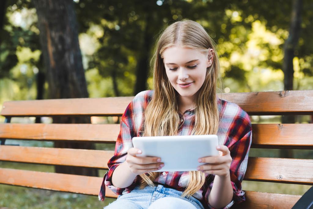 νεαρό κορίτσι σε casual ρούχα κάθεται σε ξύλινο παγκάκι στο πάρκο και χρησιμοποιώντας το ψηφιακό δισκίο - Φωτογραφία, εικόνα