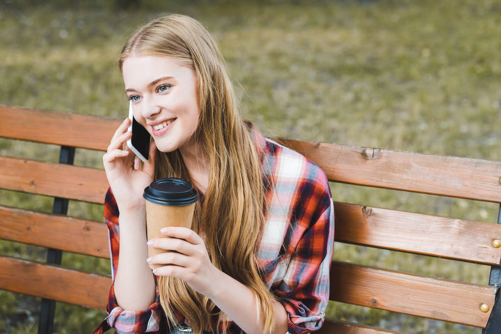 kaunis tyttö rento vaatteet tilalla paperi kahvikuppi istuessaan puinen penkki puistossa ja puhuu älypuhelin
 - Valokuva, kuva