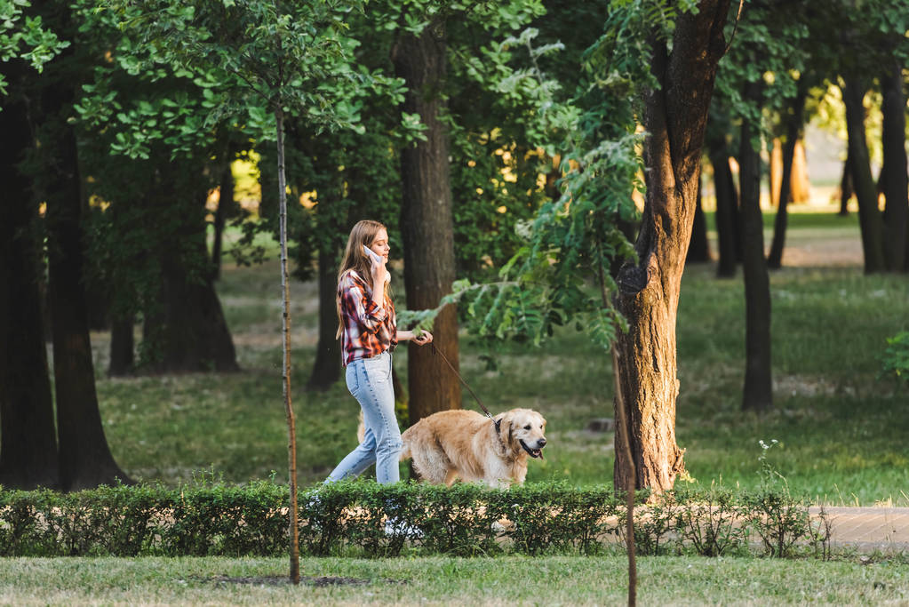 volledige lengte weergave van mooi meisje in Casual kleding wakker in Park met Golden Retriever en praten op smartphone - Foto, afbeelding