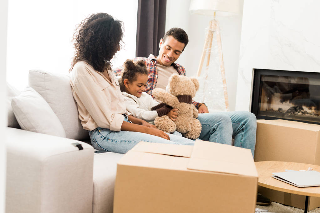 Afrikanisch-amerikanische Familie sitzt auf dem Sofa und lächelt, während Tochter mit Teddybär spielt und Eltern auf Kind schauen  - Foto, Bild
