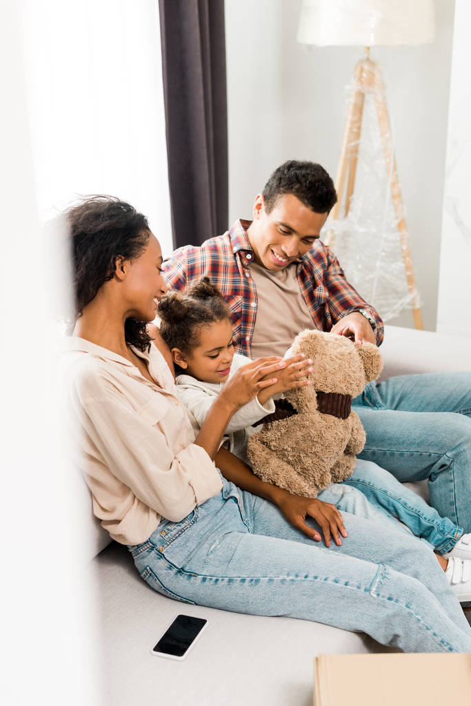 selektywne skupienie Afryki amerykańskiej rodziny siedzi na kanapie i uśmiechając się podczas gdy córka grając z misia i rodzice patrząc na dziecko  - Zdjęcie, obraz