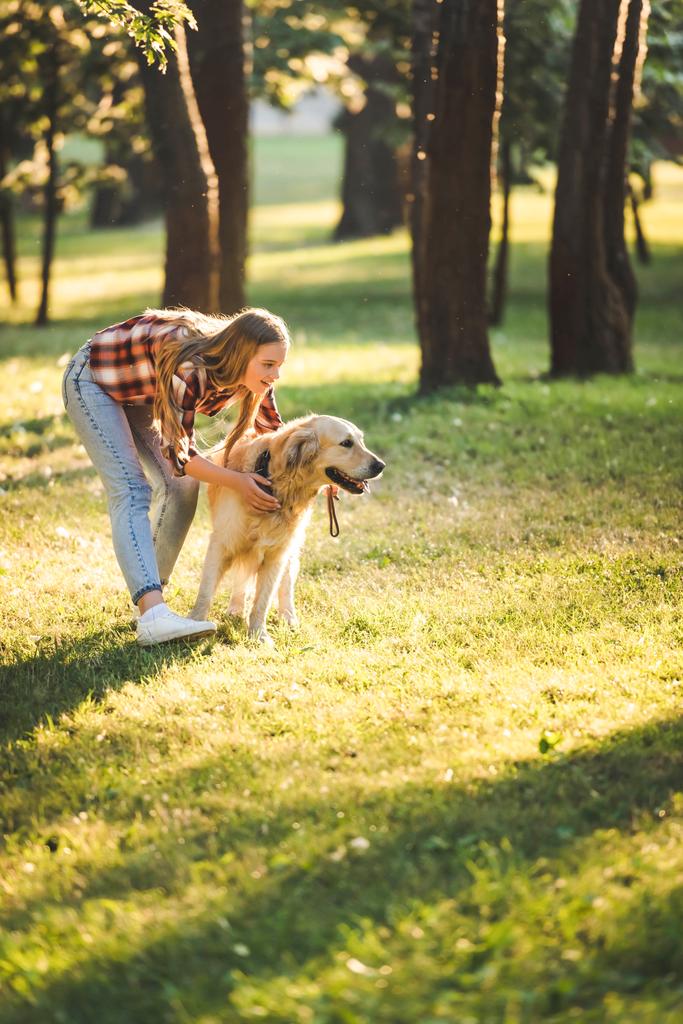 full length view of beautiful young girl in casual clothes hugging golden retriever while standing on meadow in sunlight - Photo, Image