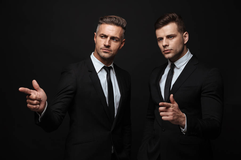 Portrét opravdu hezkého dvou podnikatelů oblečených do formálního obleku ukazujícími prsty a s pohledem stranou - Fotografie, Obrázek