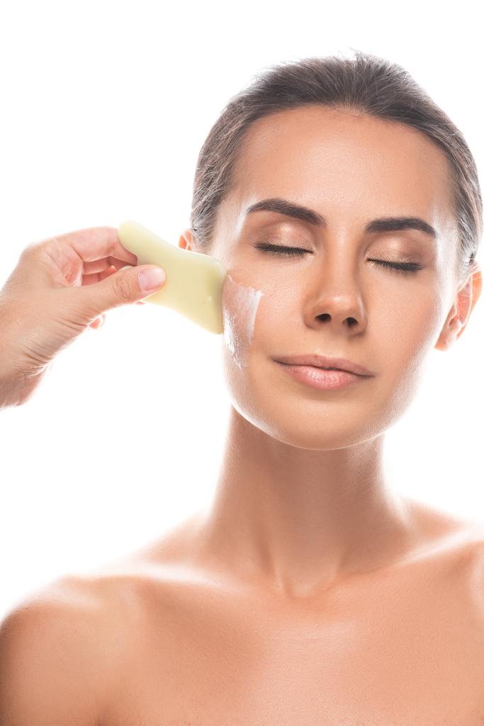vista parcial del cosmetólogo aplicando crema facial y utilizando raspador de gua sha aislado en blanco
 - Foto, imagen