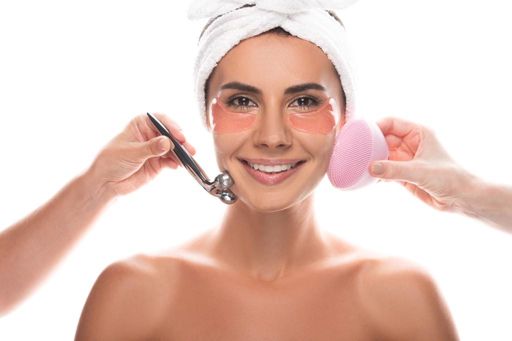 обрезанный вид косметологов с помощью щетки для очищения лица и массажистки для лица и улыбающейся женщины с повязками на глазах, изолированными на белом
 - Фото, изображение