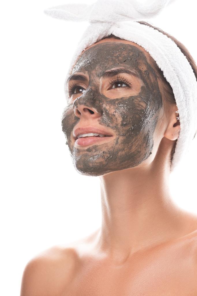 naakt jonge vrouw in cosmetische haarband met Clay masker op gezicht op zoek weg met glimlach geïsoleerd op wit - Foto, afbeelding