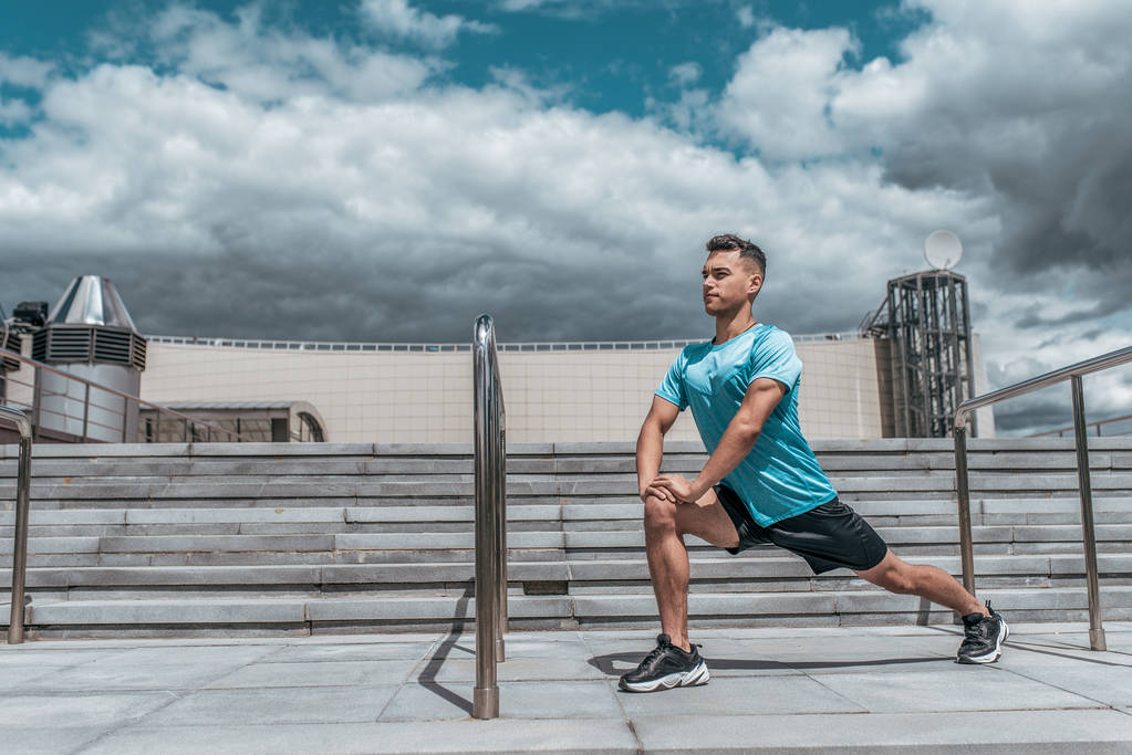 男性アスリートは、ジョギング、フィットネストレーニング、スポーツウェア、集中力と目標に焦点を当てる前に筋肉を伸ばし、夏の街を肺にします。階段の背景青い空。テキストの空き領域. - 写真・画像
