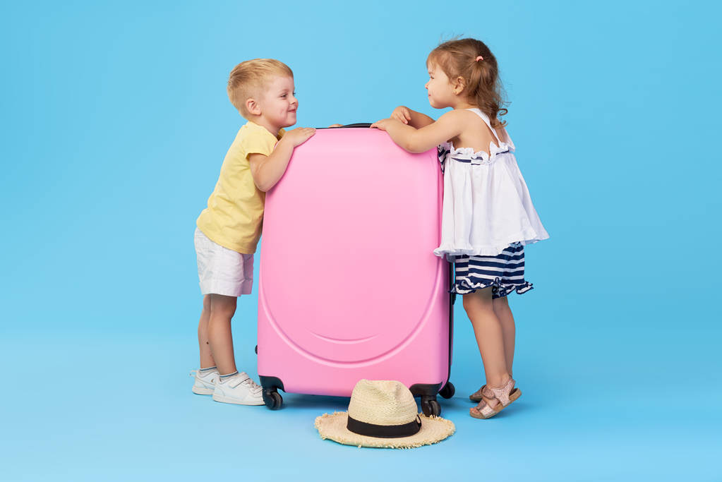 Onnelliset lapset tilalla värikäs vaaleanpunainen matkalaukku valmis kesälomalle. Nuoret matkustajat. Pieni tyttö ja poika, sisko ja veli, hauskaa eristetty sinisellä taustalla
 - Valokuva, kuva