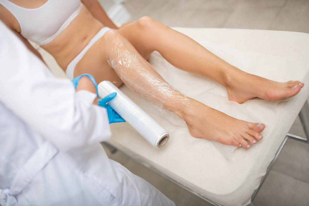 Kosmetikerin deckt Bein einer Kundin mit Frischhaltefolie ab. - Foto, Bild