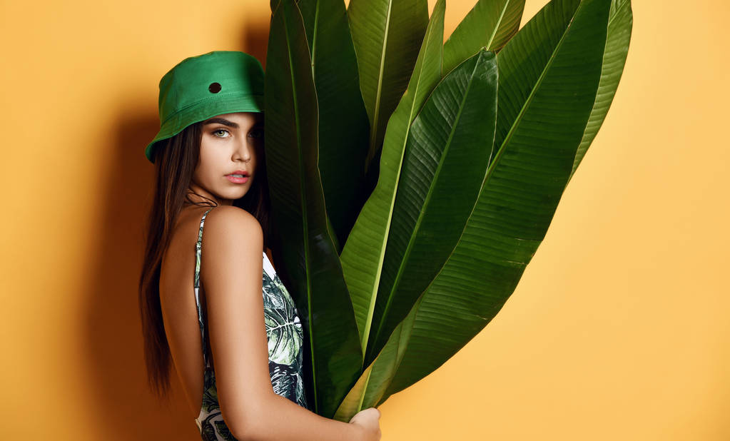 Menina de moda jovem bonita com pele perfeita em chapéu verde segurar folha de banana tropical em mãos e tampas
 - Foto, Imagem