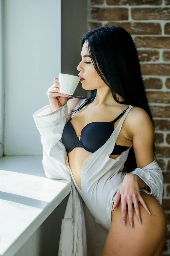 Rano kawa poprawia jej nastrój. Zmysłowa dziewczyna delektująca się aromatyczną poranną kawą. Seksowna kobieta pijąca zdrowego poranka przy oknie. Kawa budzi ją rano. - Zdjęcie, obraz