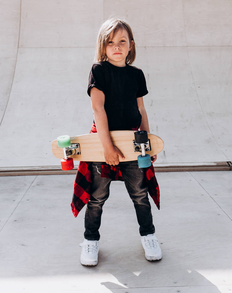 El niño vestido con la ropa casual con monopatín en la mano se para en un parque de skate junto al tobogán en el día soleado afuera.
 - Foto, Imagen
