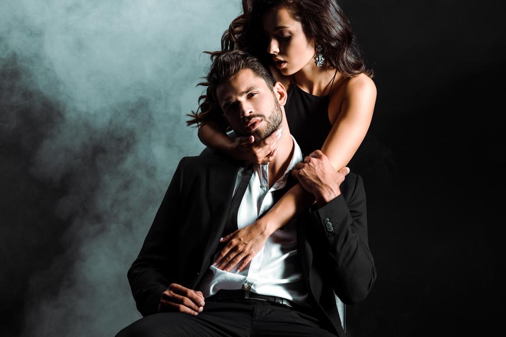 passionate young woman standing and hugging handsome bearded man on black with smoke  - Photo, Image