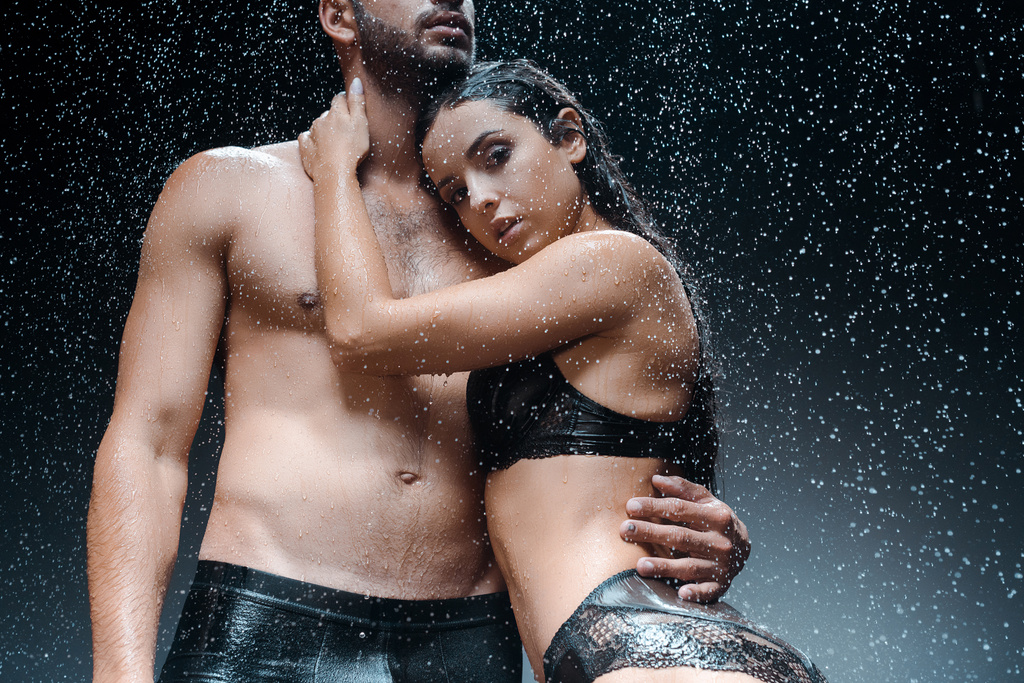 περικομμένη όψη του υγρού άντρα χωρίς μπλούζα στέκεται με το κορίτσι κάτω από σταγόνες βροχής στο μαύρο  - Φωτογραφία, εικόνα