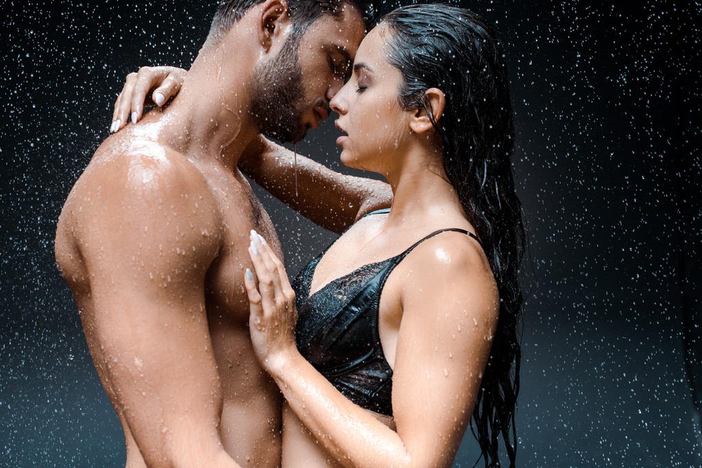 黒の雨滴の下でシャツなしと濡れ男を抱きしめる魅力的な女性  - 写真・画像