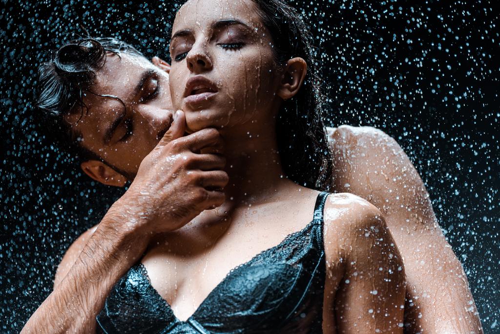 niski kąt widzenia tortu i mokrego człowieka dotykania twarzy atrakcyjnej dziewczyny z zamkniętymi oczami pod deszczem na czarno  - Zdjęcie, obraz