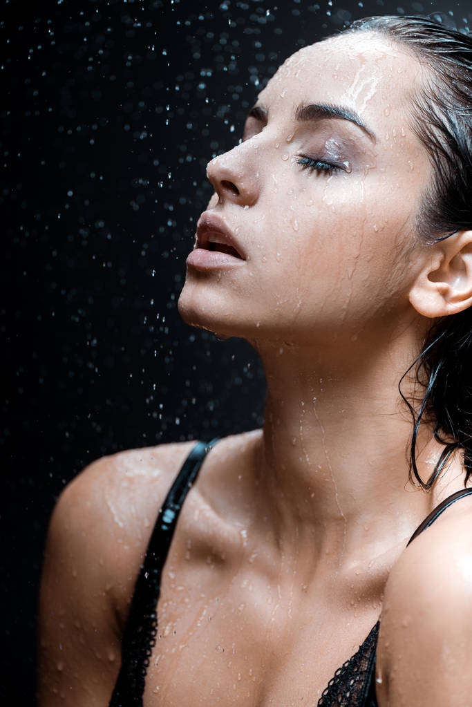 attraktive junge Frau mit geschlossenen Augen unter Regentropfen auf Schwarz  - Foto, Bild