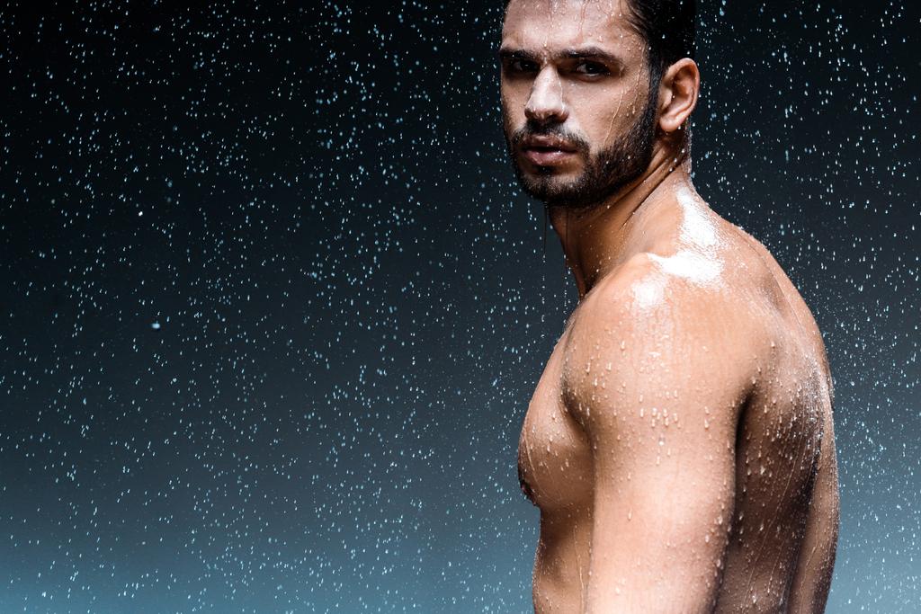 wet man looking at camera while standing under raindrops on black  - Photo, Image