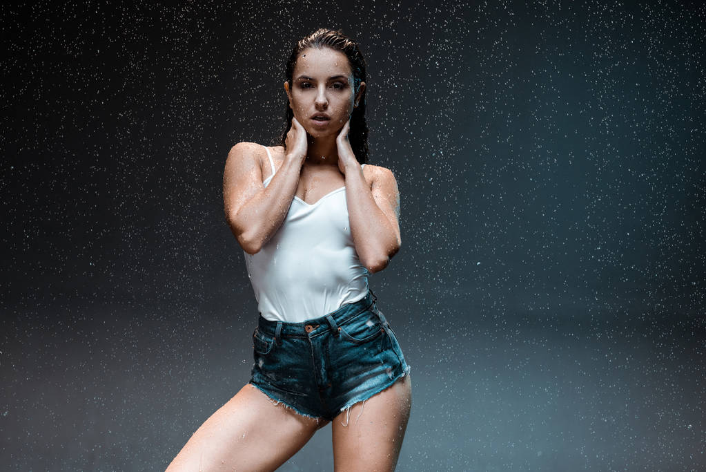 sexy junge Frau steht und schaut in die Kamera unter Regentropfen auf schwarz  - Foto, Bild
