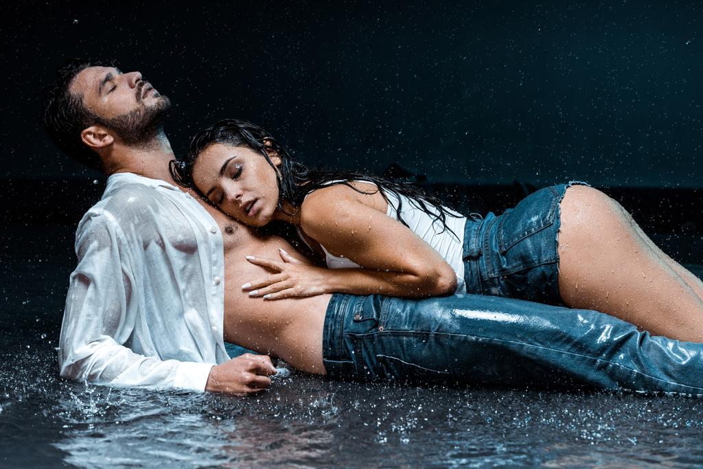 σέξι και υγρή γυναίκα που κείτεται με όμορφο φίλο κάτω από σταγόνες βροχής στο μαύρο  - Φωτογραφία, εικόνα